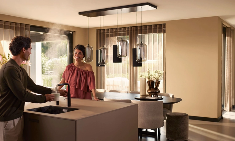 Man en vrouw in moderne keuken, met kokendwaterkraan en sfeervolle verlichting boven de eettafel.